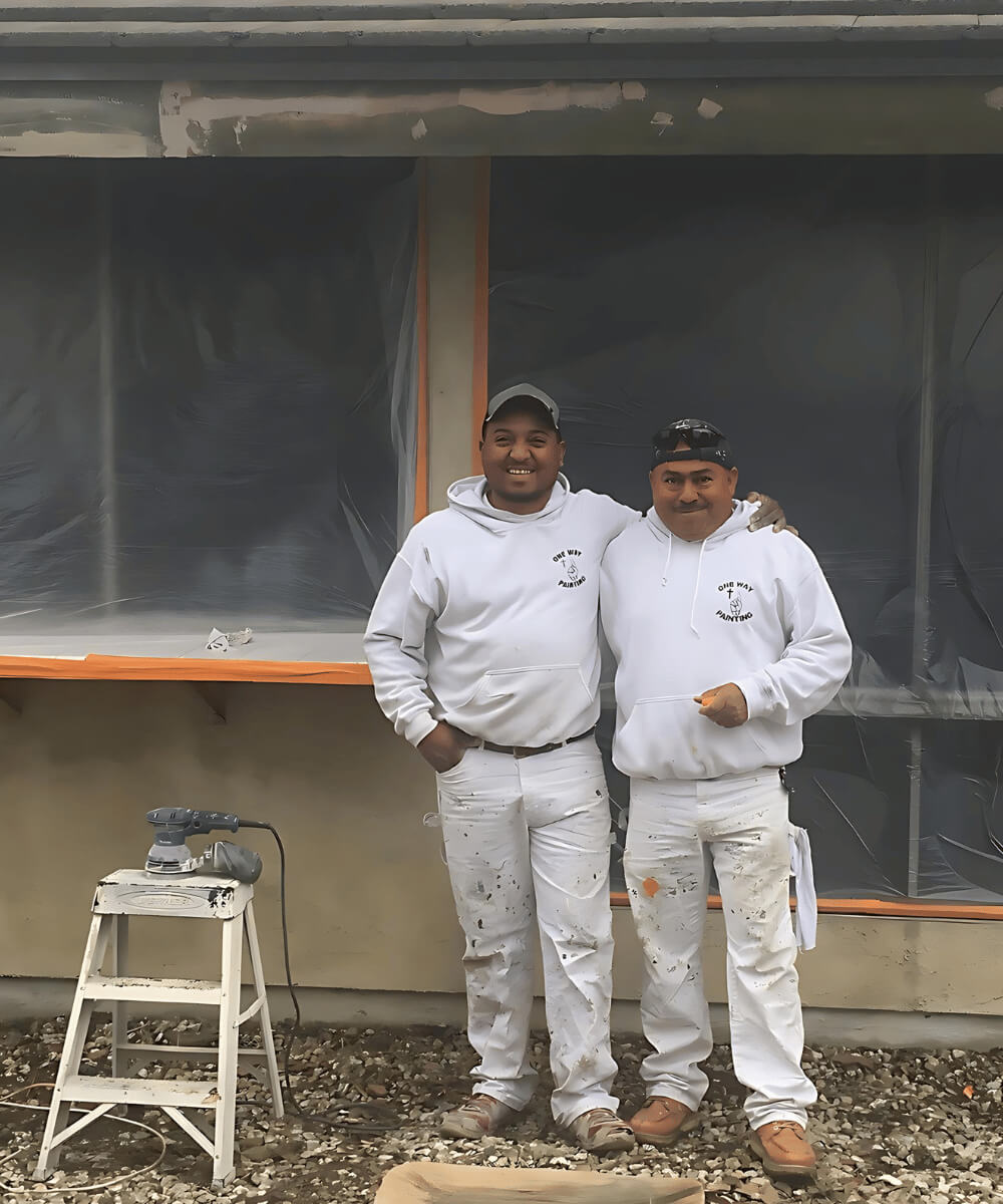 Two One Way Painting house painters smiling and posing for a photo in front of a house with plastic coverings over the windows.