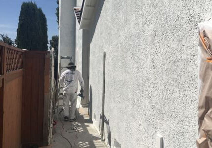 Painter prepping the stucco on the exterior wall of a house for painting, working in a narrow space between the house and a fence.